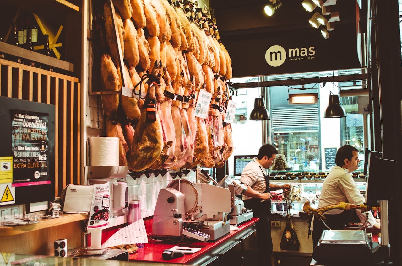 Productos cárnicos en Sort. A bustling Spanish ham shop with hanging cured meats and two men serving customers.